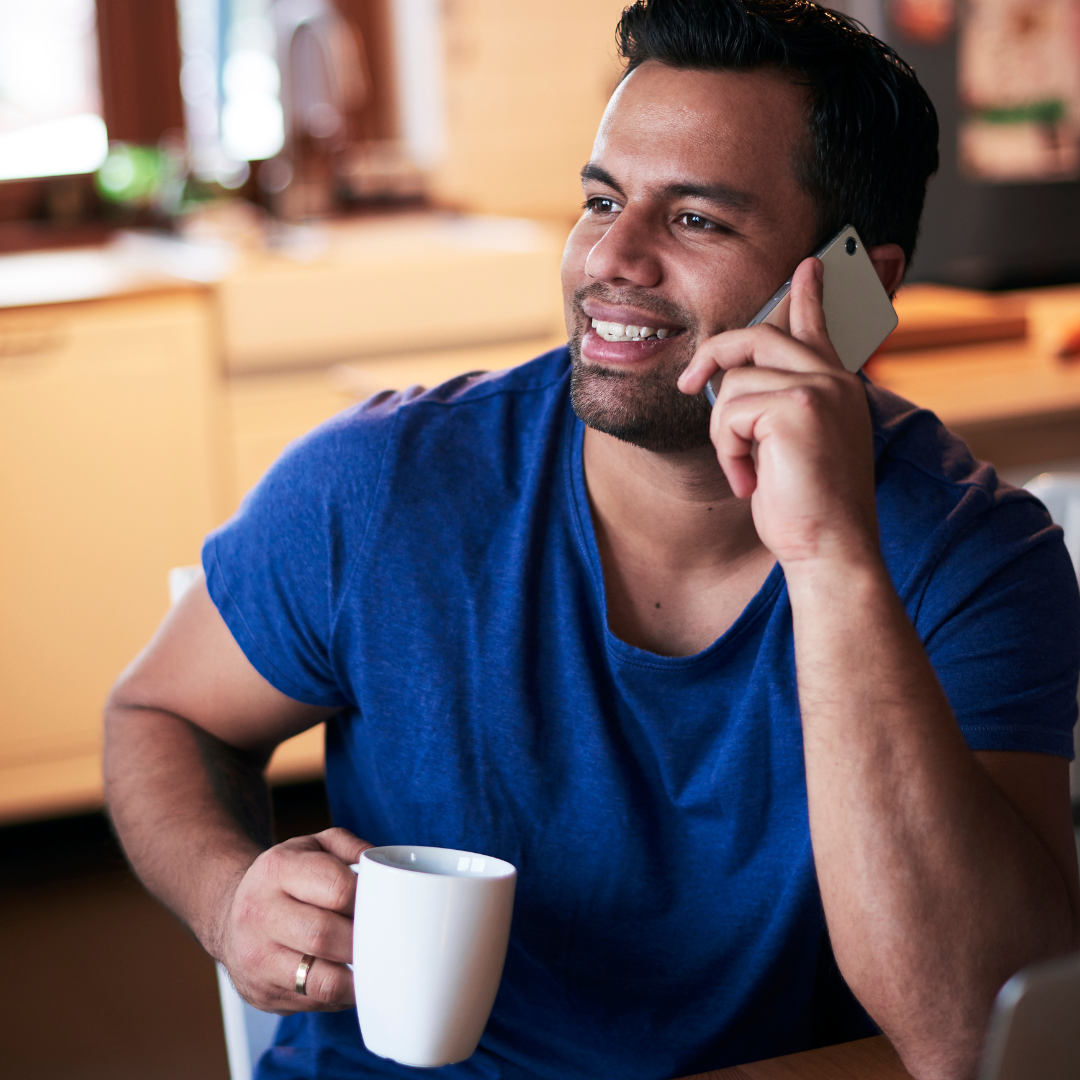Young man in blue T-shirt talking on the phone about Dutch lessons in Amsterdam, while holding a cup of coffee in his right hand.