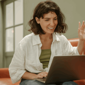 Woman in her living room with a laptop on her lap, waving friendly to her private Dutch tutor.