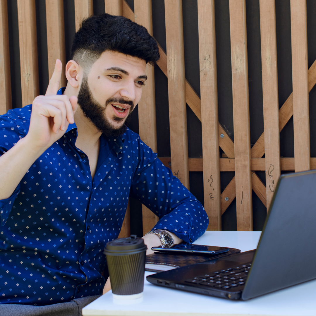 Man in a dark blue shirt taking online Dutch Sprint lessons from his garden. He speaks enthusiastically to his tutor. There is a cardboard cup of takeaway coffee on the table.