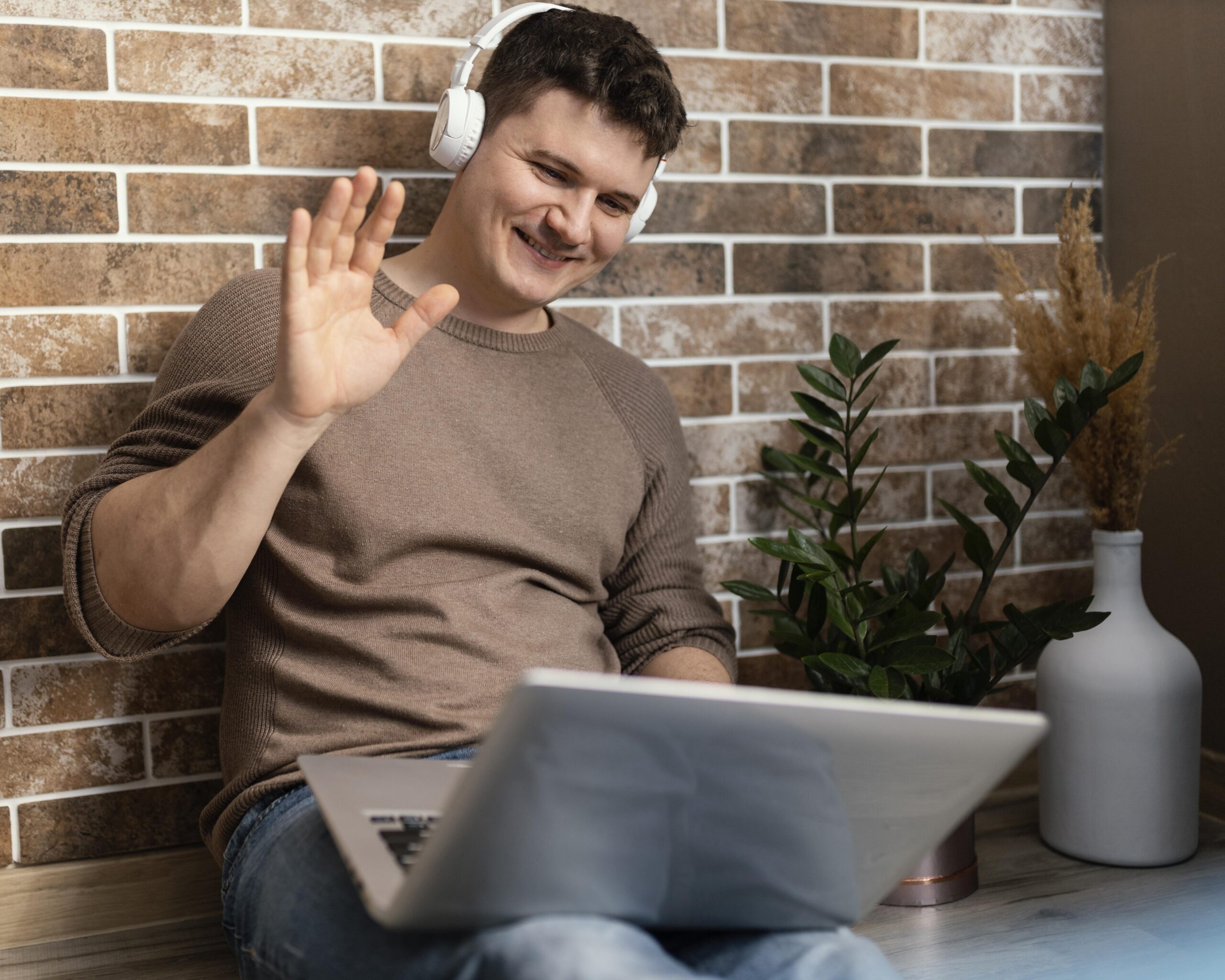 Young man in brown sweater wearing headphones and waving at his laptop screen to greet his Dutch tutor who helps him master essential Dutch in one month.