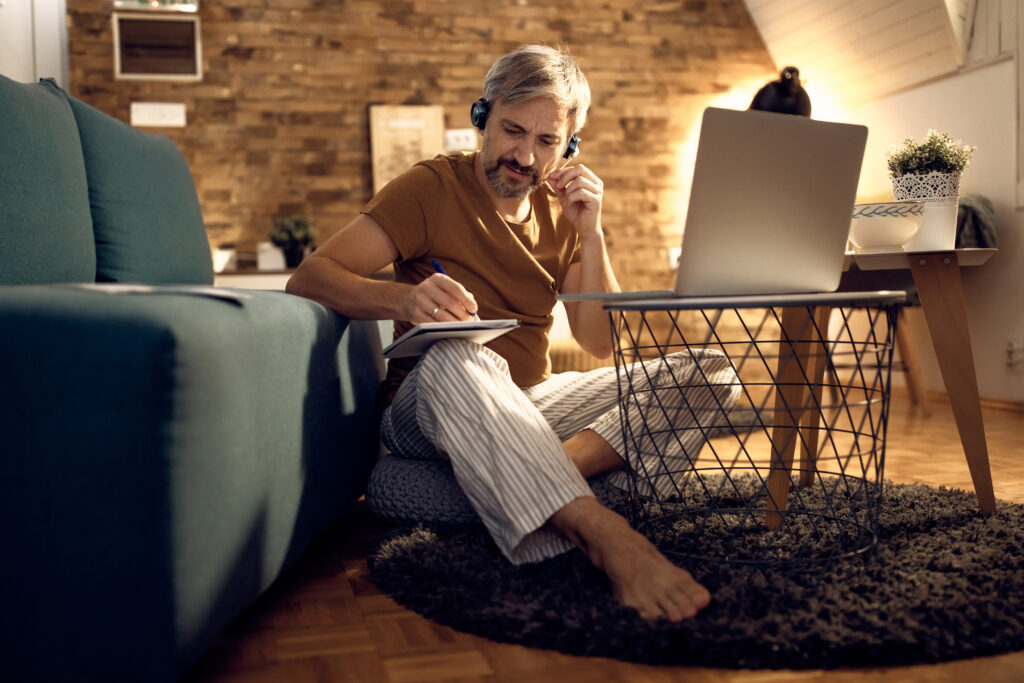A man in pyjamas, sitting on the floor of his living room, studying Dutch while wearing headphones and taking notes.