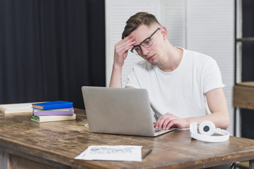 A young expat sitting at a wooden desk, looking at his laptop in despair, rubbing his forehead with his hand. Bite-sized and structured materials typically work better than text-heavy grammar books.